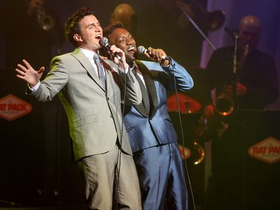 Two men, one in a grey suit and the other in a blue suit, both sing together and lean back slightly with their eyes closed