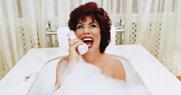 Ruby Wax in a bubble bath with net curtains behind her laughing on the phone