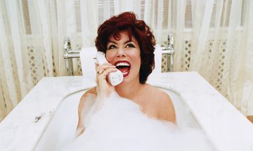 Ruby Wax in a bubble bath with net curtains behind her laughing on the phone