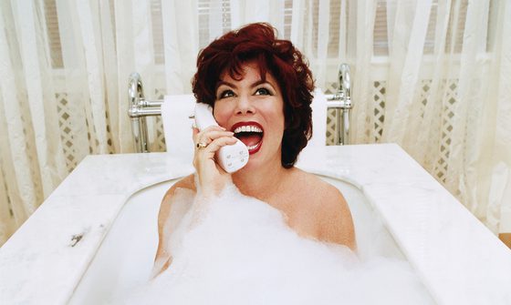 Ruby Wax in a bubble bath with net curtains behind her laughing on the phone