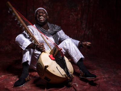 Seckou sits on the floor while wearing an all white outfit, he laughs while holding a Kora