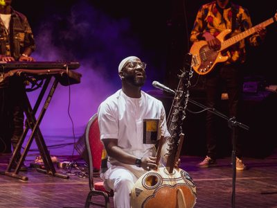 Seckou wears a white t-shirt and trousers, he sits on stage and sings into a microphone while playing the Kora, behind him is a pianist and a guitarist