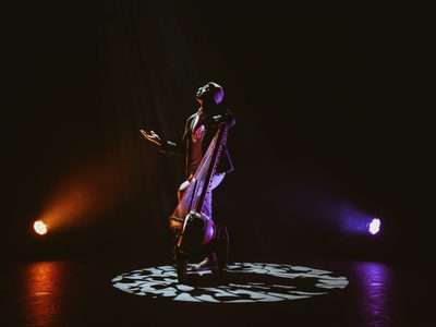 Seckou stands centre stage, lit by a spotlight, he looks up with one hand out to his side while he holds the Kora