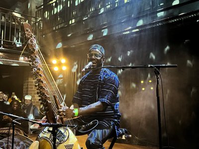 Seckou, black man in a suit sits on stage at a Jazz Cafe and sings into a microphone while playing the Kora, lit by a disco ball