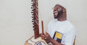 Seckou, a young black man in a white t-shirt sings while playing the Kora