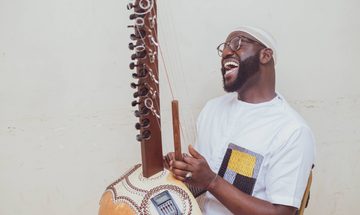 Seckou, a young black man in a white t-shirt sings while playing the Kora