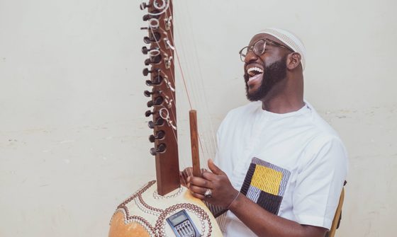 Seckou, a young black man in a white t-shirt sings while playing the Kora