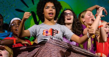 A group of children in brightly coloured clothes all shouting on stage
