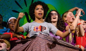 A group of children in brightly coloured clothes all shouting on stage