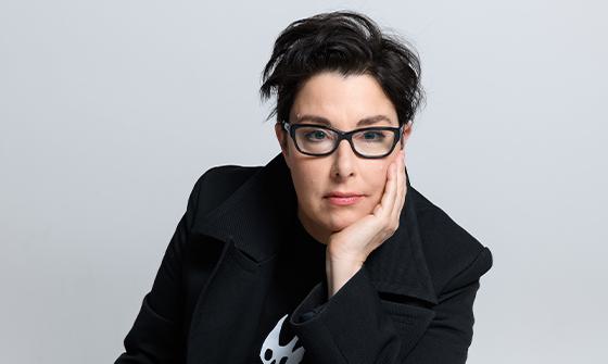 Sue Perkins with short dark hair, wearing glasses and a black coat, resting her face on her hand while looking at the camera