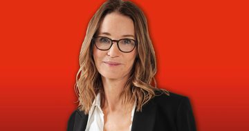 Susie Dent in a white shirt and black blazer stands against a bright red background