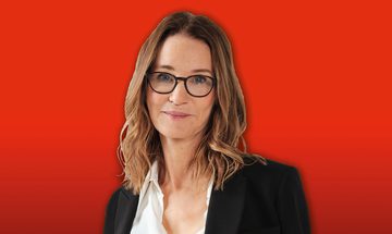 Susie Dent in a white shirt and black blazer stands against a bright red background
