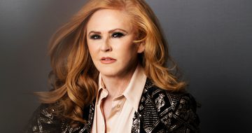 A glamorous red haired woman sits posed in a blush pink stain shirt and a black knit cardigan