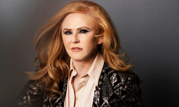 A glamorous red haired woman sits posed in a blush pink stain shirt and a black knit cardigan