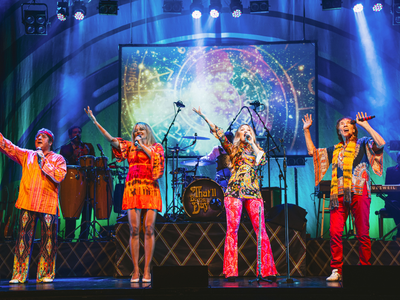 Four singers, two men, two women stand centre stage all singing into hand held microphones wearing 60s themed outfits