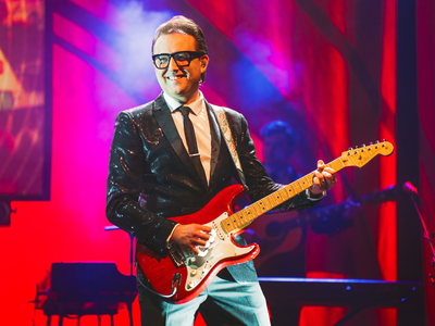A man in jeans and a black suit jacket smiles as he plays a red guitar