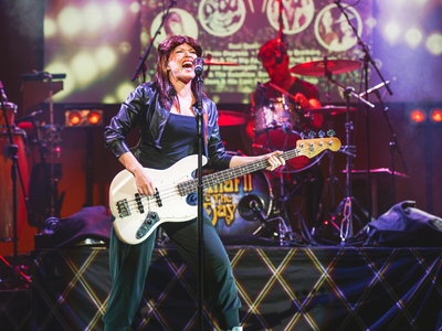 A woman with brown hair and an all black outfit stands on stand singing while playing a white guitar