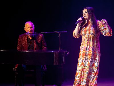 On a dimly lit stand a woman in an orange patterned dress sings into a handheld microphone while an older man plays the piano next to her, watching her sing