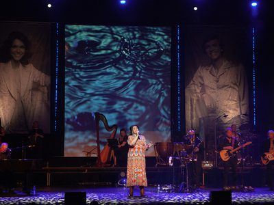 On a dimly lit stage a woman in an orange floral dress sings in the spotlight, behind her is a large band