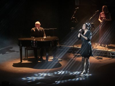 Lit by a spotlight a woman in a black dress sings as a man, also under a spotlight, plays the piano