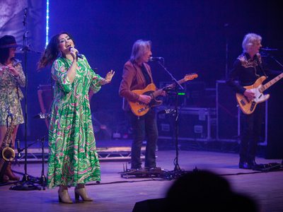 A woman in a bright green patterned dress swings on the spot as she sings, behind her are two guitar players and a woman playing the flute
