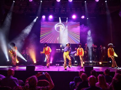 Five performers dressed in retro clothing sing and dance on stage under purple lighting