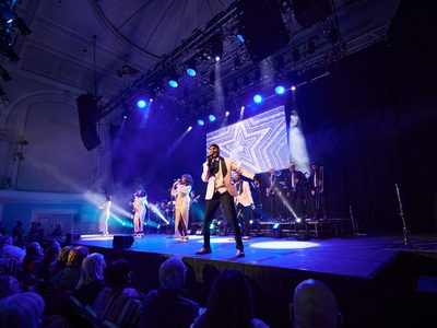 Four performers all dressed in white stand in a row across the blue lit stage and sing