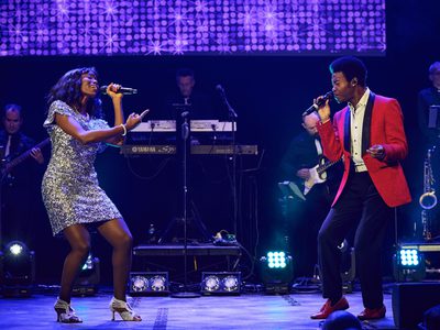A black woman in a leopard print mini dress and a black man in a red and black suit face each other and sing