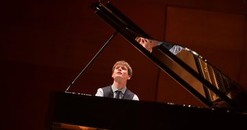 A photo taken behind a piano of Thomas Luke performing in a shirt, waistcoat, and tie while looking towards the top left of the photo