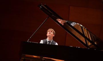 A photo taken behind a piano of Thomas Luke performing in a shirt, waistcoat, and tie while looking towards the top left of the photo