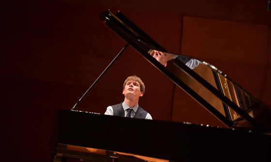 A photo taken behind a piano of Thomas Luke performing in a shirt, waistcoat, and tie while looking towards the top left of the photo
