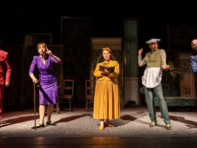 Five people on stage under spotlights: a man in a red suit and tie, a woman in a purple pencil skirt and blazer with a walking stick, a woman in a yellow jacket and long skirt holding a small book, a man in a green suit with a small white apron and large chef hat, a man in a blue suit