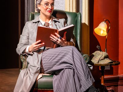 A woman dressed in a grey suit and grey wig with round glasses sat in an armchair smiling at the camera and holding a large book