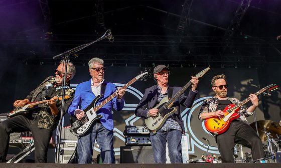 Four men wearing sunglasses play electric guitars on an outdoor concert stage.