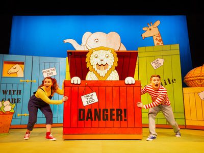 A man and woman either side of a large red crate with a lion jumping out. Behind are other colourful crates with a camel, elephant, and giraffe inside