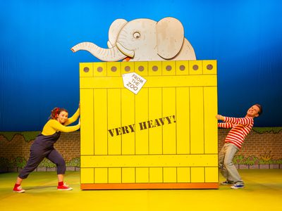 A man and a woman pushing and pulling on either side of a large yellow crate with an elephant sticking out the top
