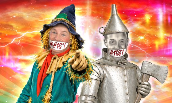 Bobby Davro dressed as a scarecrow and Stevi Ritchie dressed as the Tin Man standing together smiling. Bobby is pointing at the camera and Stevi is holding an axe. They both have white cloths over their mouths with red comic expletives