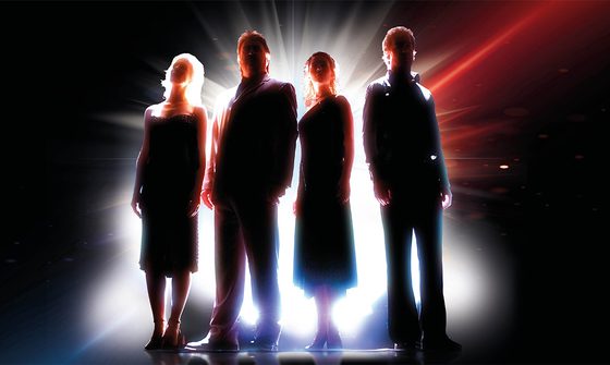 Four people in formal black attire stand silhouetted against dramatic backlighting