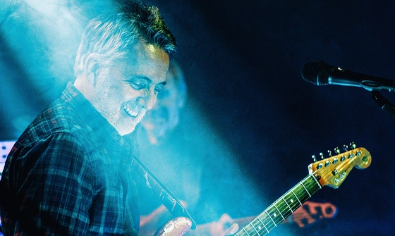 An older white man in a plaid shirt under stage lights holding and electric guitar in front of a microphone smiling