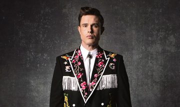 Ed Gamble stood in a black cowboy jacket embroidered with flowers and a large E and G with white accents and fringe. He is also wearing a white shirt with black bolo tie. He is stood in front of a grey wall with a blank expression
