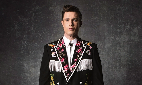 Ed Gamble stood in a black cowboy jacket embroidered with flowers and a large E and G with white accents and fringe. He is also wearing a white shirt with black bolo tie. He is stood in front of a grey wall with a blank expression