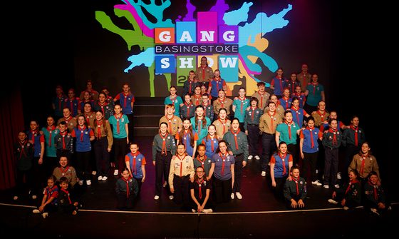 A large group of Scouts, Cubs, and Beavers, stood on stage in their uniforms with a brightly coloured logo for the Gang Show behind them