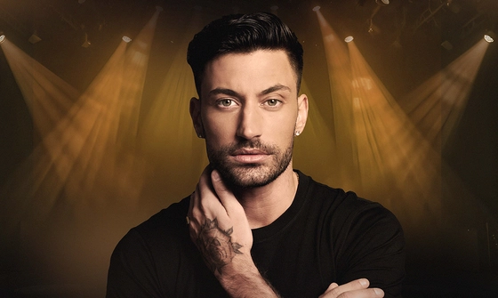 Giovanni, a tanned Italian man with dark quiffed hair and a stubbly beard in a black t-shirt and diamond earrings. His left hand is on the right side of his neck