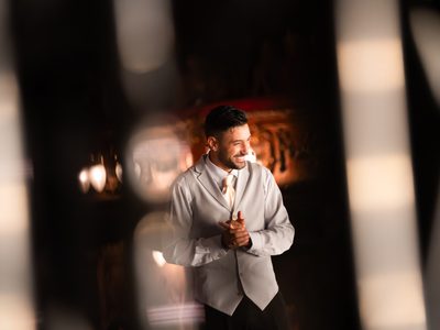 Giovanni Pernice smiles while facing to the side in a dimly lit room, wearing a silver waistcoat, white shirt and orange tie, with his hands clasped together.