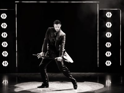 A black-and-white image of Giovanni Pernice dancing in a black suit while holding a baton, with stage lights behind him.