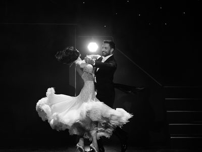 A black-and-white image of Giovanni Pernice smiling while dancing with a woman in a white fur-lined dress on stage, with steps and stage lighting behind them.
