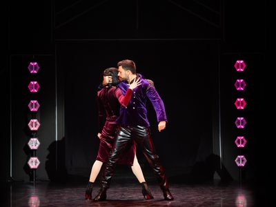 Giovanni Pernice, wearing a purple velvet blazer, stands with a female dancer in a burgundy velvet dress. She faces away from the audience while he faces forward, looking towards her and holding her hair as her hand rests on his chest, with purple and pink stage lights behind them.
