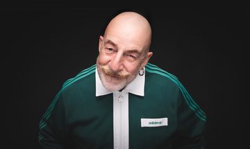 Jeff Innocent, an older bald white man with white and ginger beard and moustache, large hoop earring, and a green adidas tracksuit jacket. He's leaning forward and smiling on a black background