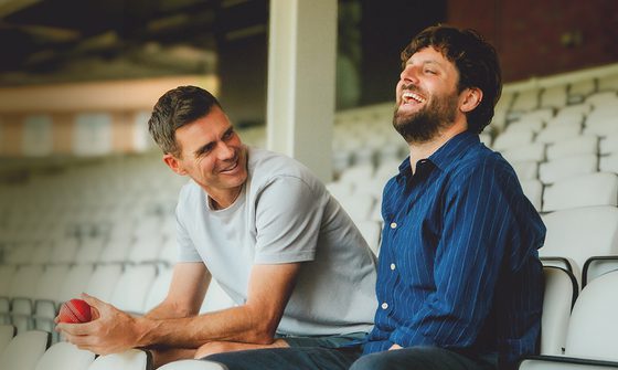 Two white men sat in the stands at a sports ground. One is in a white t-shirt holding a cricket ball leaning forward and smiling at the man next to him who's in blue button up shirt laughing with his head back.