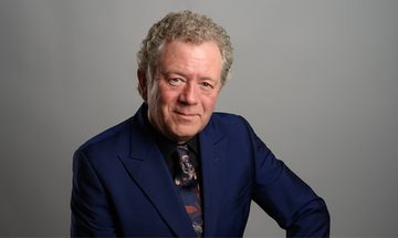Jon Culshaw, and older white man with short curly grey hair in a bule suit, black shirt, and colourful tie, softly smiling at the camera